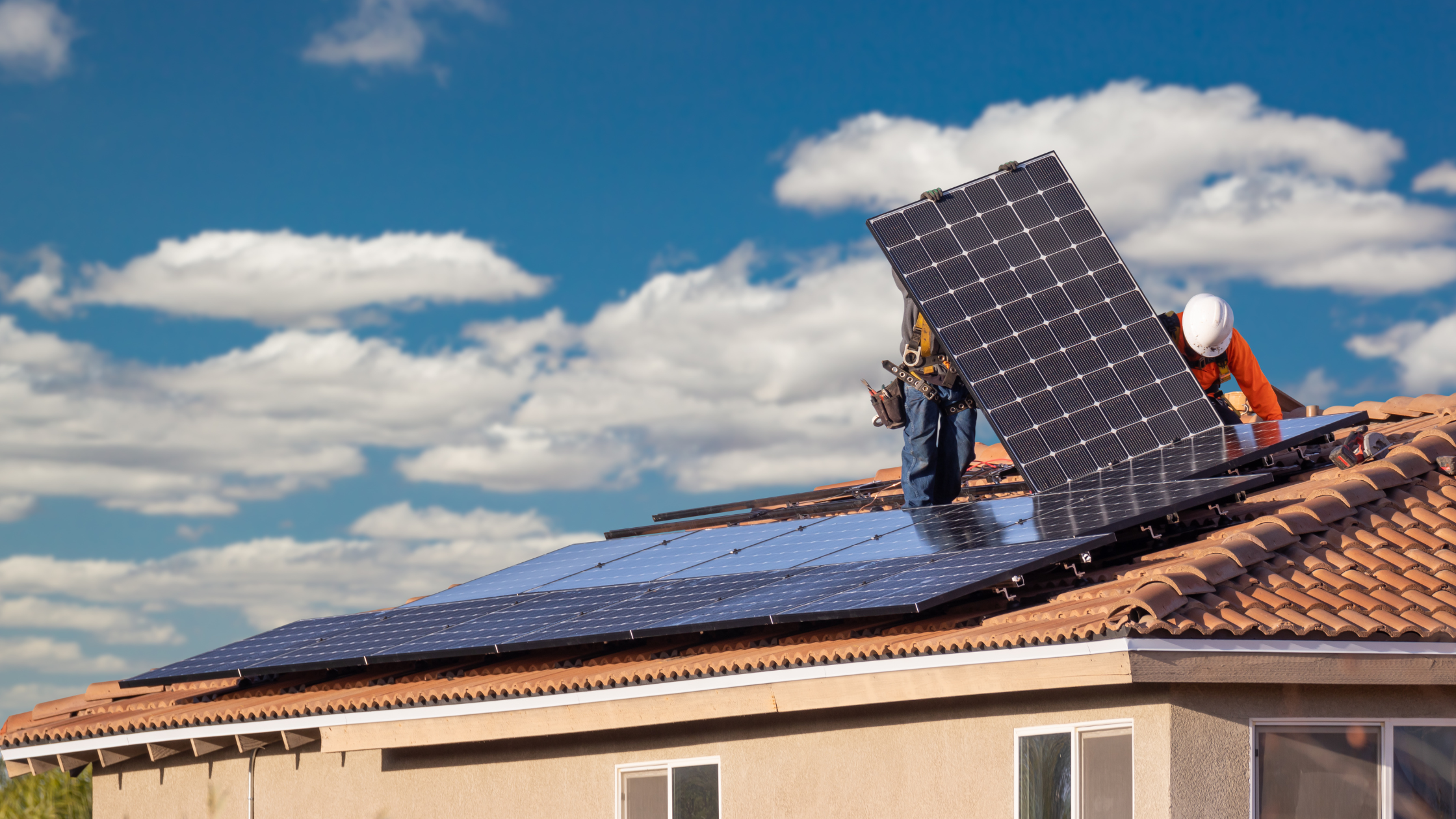 Two workers wearing safety gear install solar panels on the roof of a stucco house under a bright blue sky with scattered clouds.