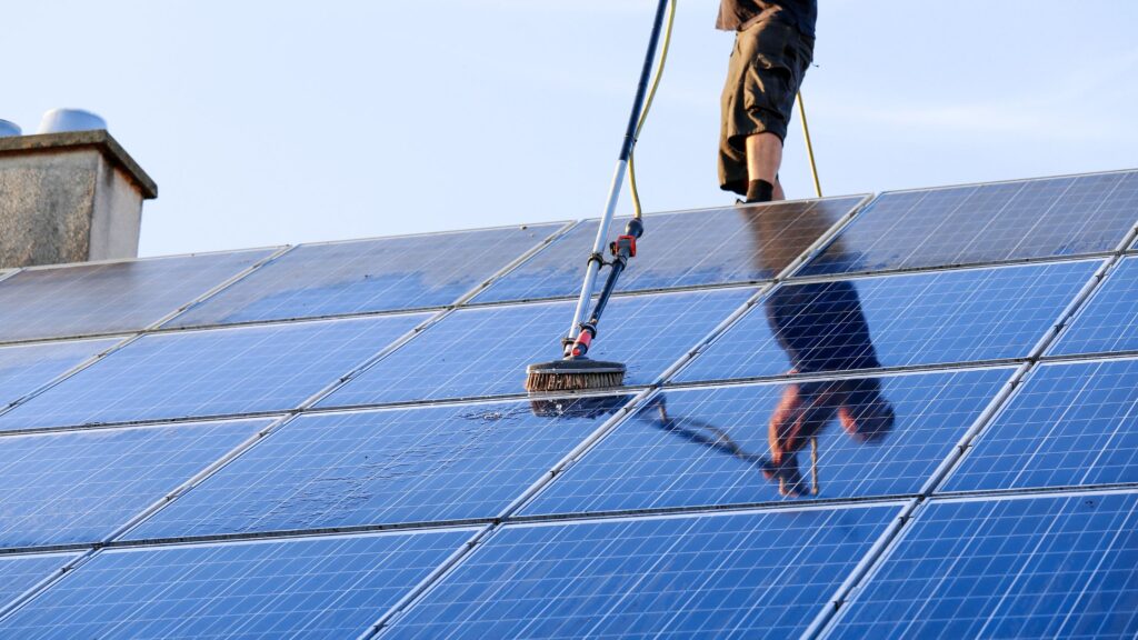 Technician cleaning rooftop solar panels with a telescoping brush to improve solar energy production and system efficiency before winter.